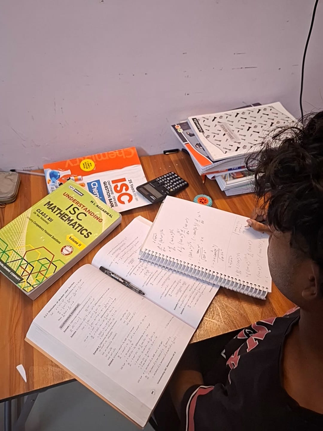 Student writing calculus notes at a wooden desk with ISC Class XII Mathematics and Chemistry textbooks and a scientific calculator